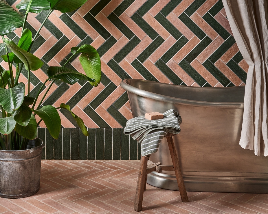 image of a bc designs boat bath in copper in front of a herringbone patterned green and beige wall with plant and terracotta herringbone floor