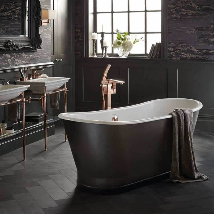 Lifestyle image of a dark themed bathroom, featuring a black painted boat bath, black chevron style floor tiles, black painted panelled walls, dark grey wallpaper and a black painted framed mirror