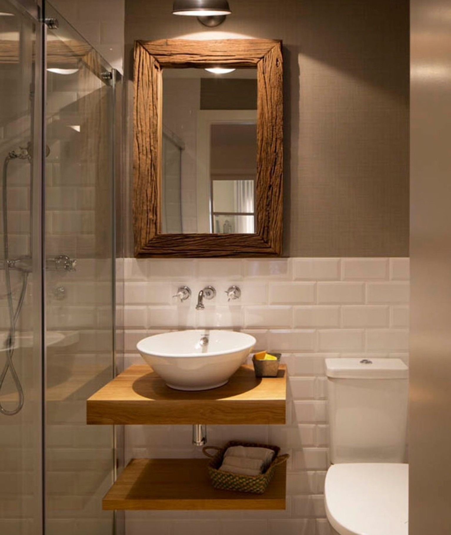 Lifestyle image of a white and brown bathroom design, featuring a wall mounted wooden shelf and countertop, a wooden framed mirror, grey-brown painted walls, white tiled walls and white ceramics 