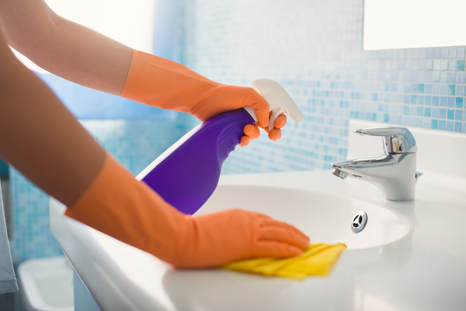 Close up image of someone wearing orange rubber gloves cleaning a basin tap using a cloth and spray bottle