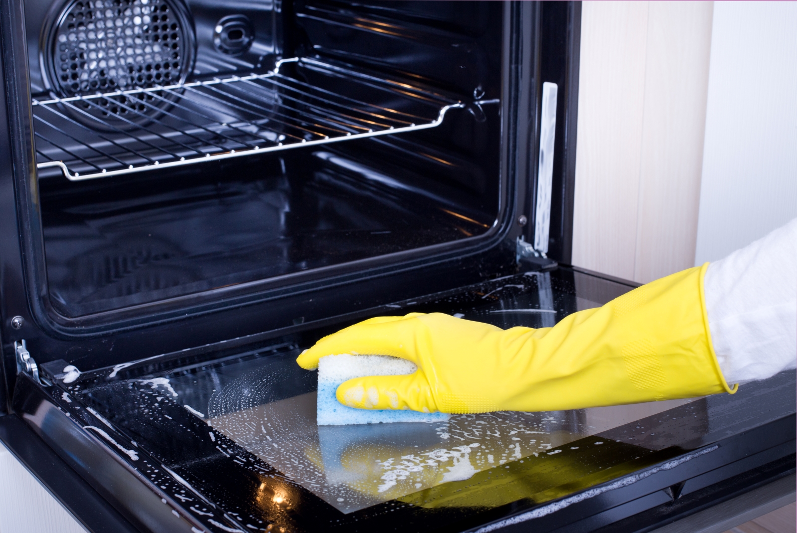 Close up image of someone wearing a yellow rubber gloves cleaning an oven with a sponge and soapy water
