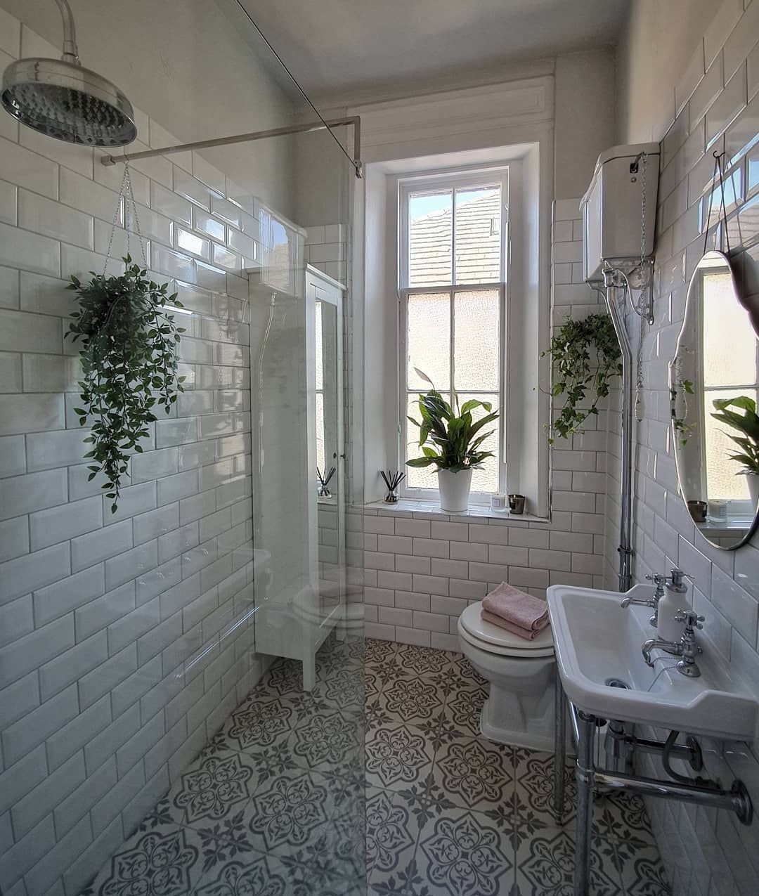 Lifestyle image of an Edwardian style bathroom, with grey patterned floor tiles, white wall tiles, a glass screened shower enclosure, a chrome washstand with a stepped basin and crosshead basin taps, an open back toilet with a high level cistern and a painted white bathroom cabinet