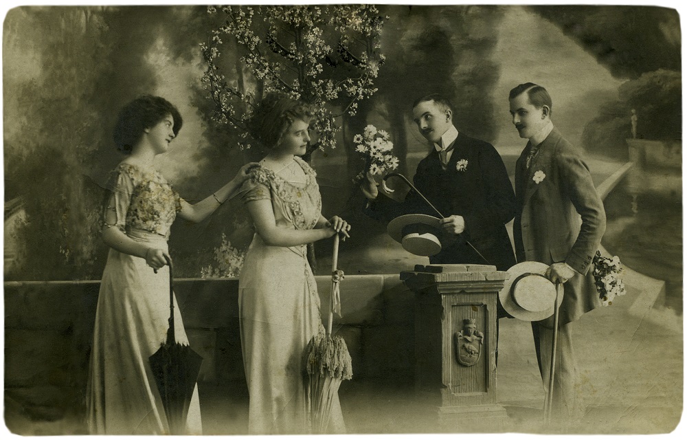 Sepia tone photograh of two Edwardian women wearing low neckline summer dresses, holding parasols, and two Edwardian men wearing linen suits, neckties, holding straw boater hats and canes