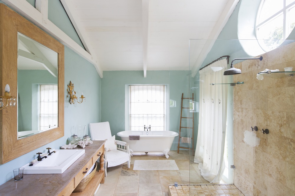 Lifestyle image of a farmhouse style bathroom, with wooden floors and turquoise walls, a walk in shower enclosure, wooden washstand, white freestanding bath with painted legs and white curtains across the windows