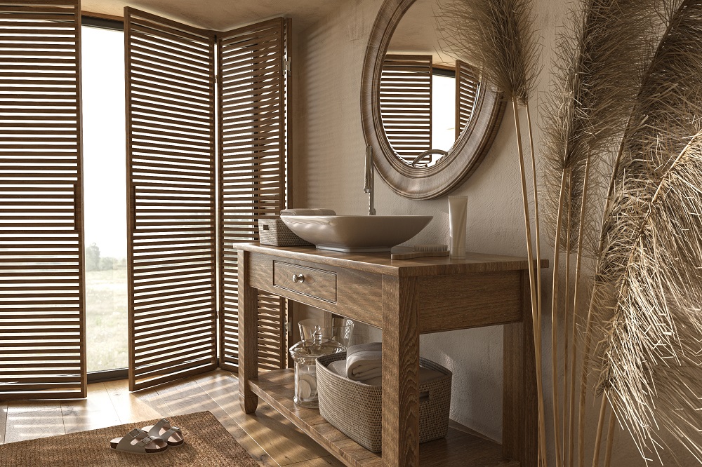 Lifestyle image of a farmhouse style bathroom, whith wooden floors and cream walls, a wooden washstand with a countertop basin, a round wooden framed window and slatted wooden doors across the windows