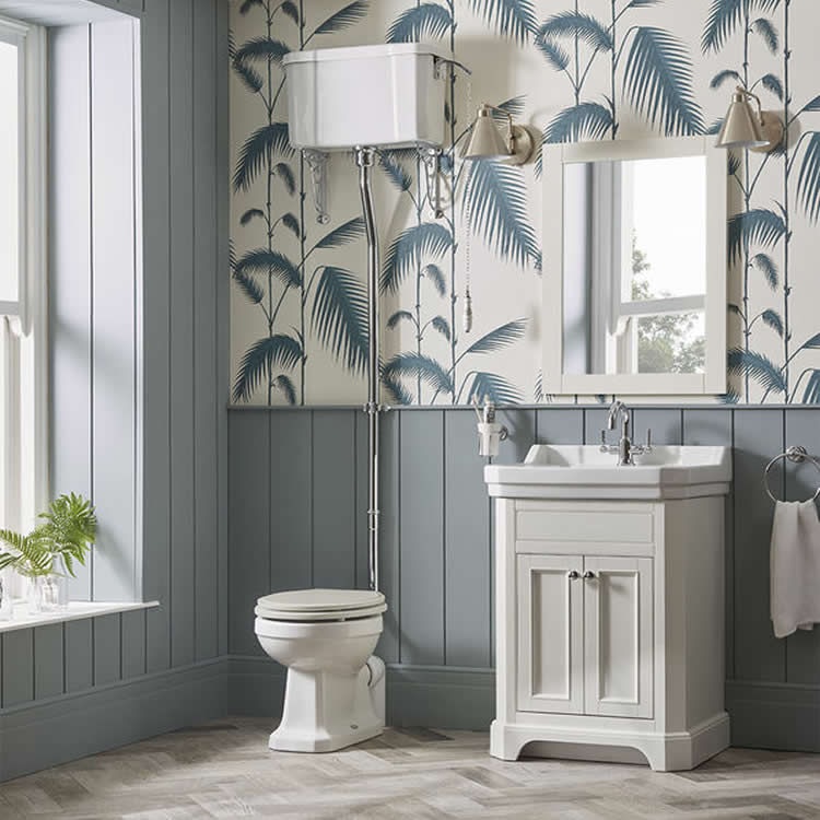 Lifestyle image of a farmhouse style bathroom, with painte blue-grey panelled walls and blue and white wall paper, a painted white wooden washbasin unit, a painted white wooden framed mirror and an open back toilet paired with a high level cistern