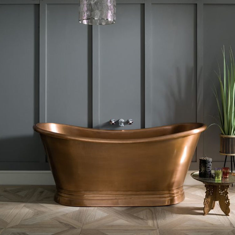 image of bc designs antique copper freestanding boat bath with roll top edges in grey walled space on wooden patterend floor with small table and spiky plant in corner of room