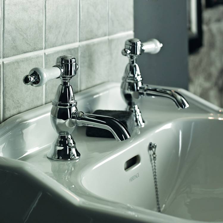close up photograph of heritage glastonbury chrome white lever pillar taps mounted on heritage basin with grey, square tiles on left and a plug and chain waste