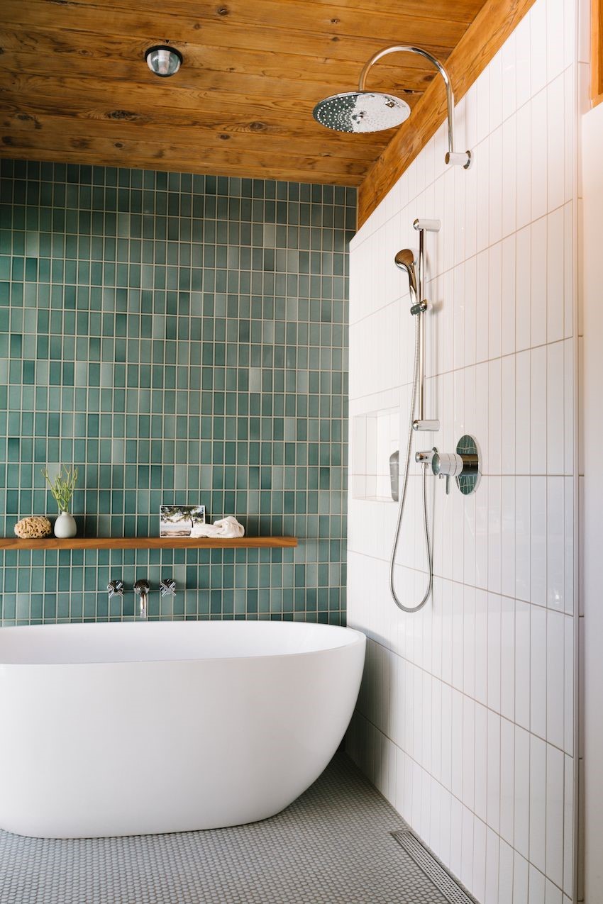image of a modern freestanding bath next to green vertical metro tiled wall with 3 tap hole chrome wall mounted tap, wooden shelf, and white tiled wall to right with shower