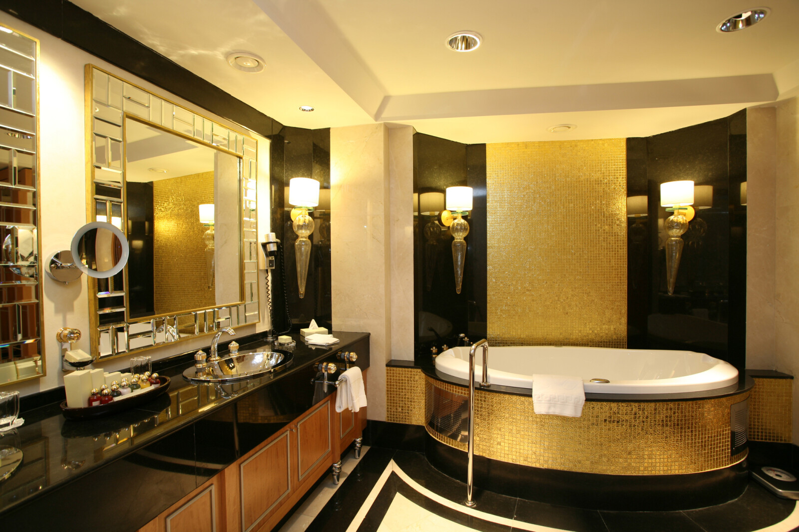 Lifestyle image of a Hotel style bathroom, featuring gold mosaic tiles on the walls and around the jacuzzi style bathtub, black and white floor tiles, cherry brown wooden cabinets with silver feet, black gloss countertop with inset chrome basin, decorative basin tap nobs and gold trim around the mirrors