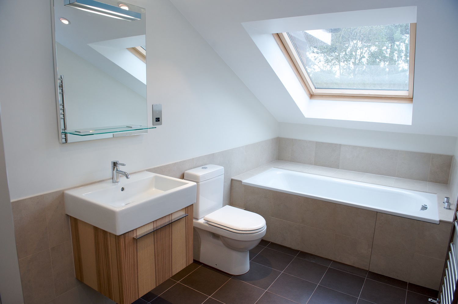 Lifestyle image of a small attic bathroom with tiled floor and walls, a built in bath below a skylight, a close coupled toilet, a wall hung wooden washbasin unit and a mirror with integrated shelf