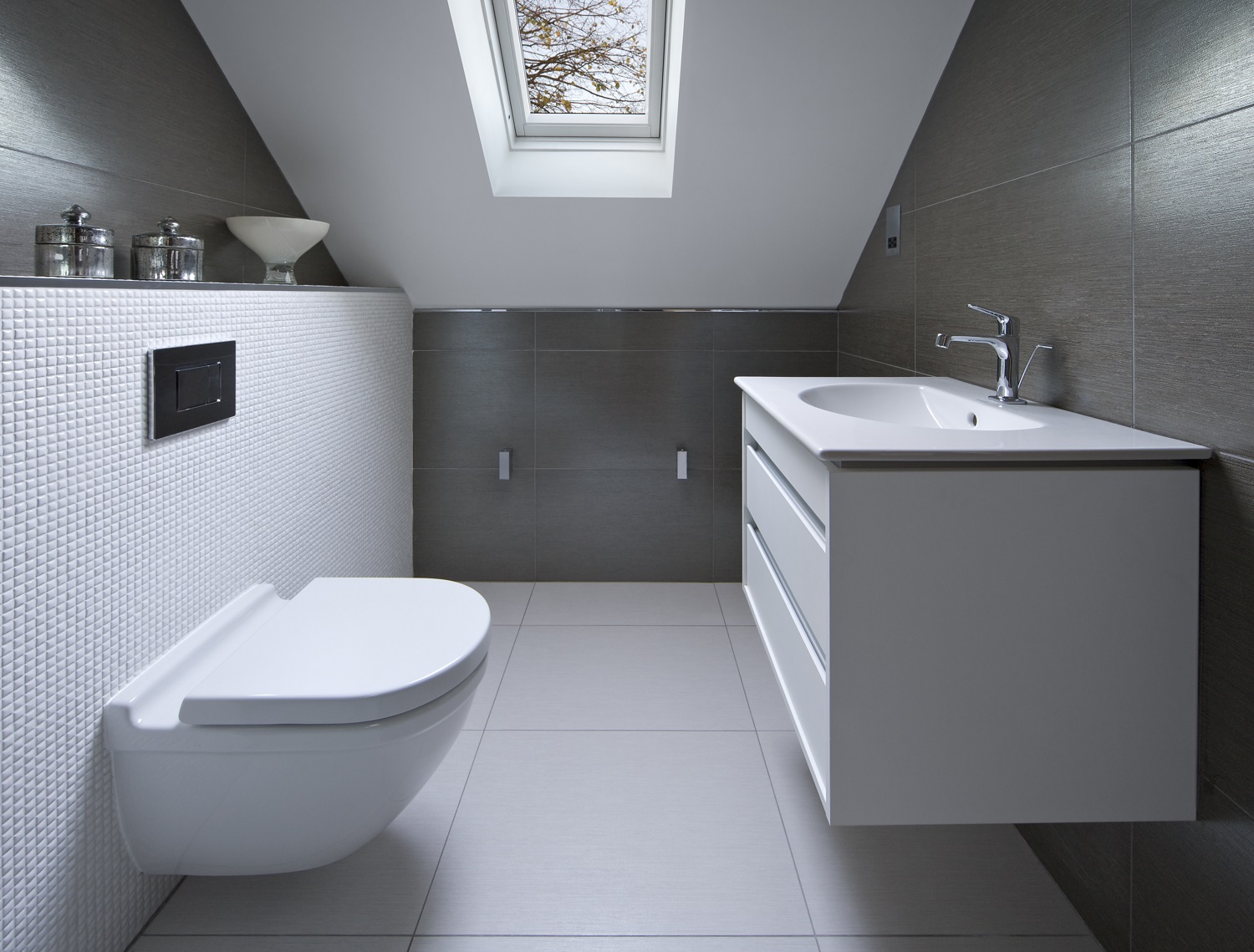 Lifestyle image of a small attic bathroom with large white floor tiles, large black and small white wall tiles, a wall hung toilet, a wall hung washbasin unit and a skylight