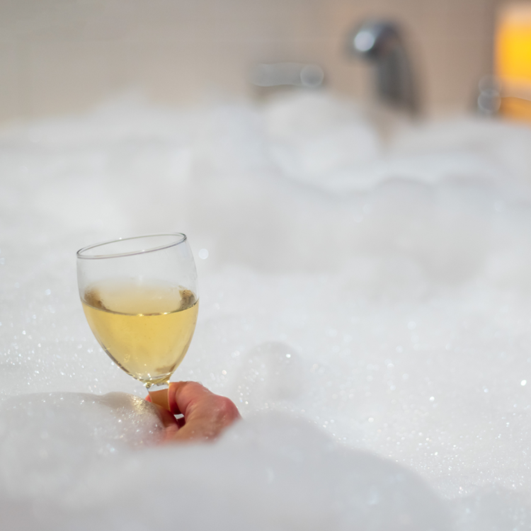 Close up image of a hand holding a glass of wine in the middle of a bubble bath