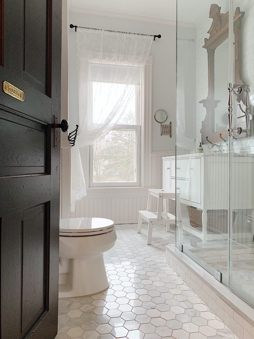 Lifestyle image of a Victorian style bathroom with white hexagonal floor tiles, a raised shower enclosure with chrome handles, a painted white vanity unit with marble worktop and a white lace curtain across the window