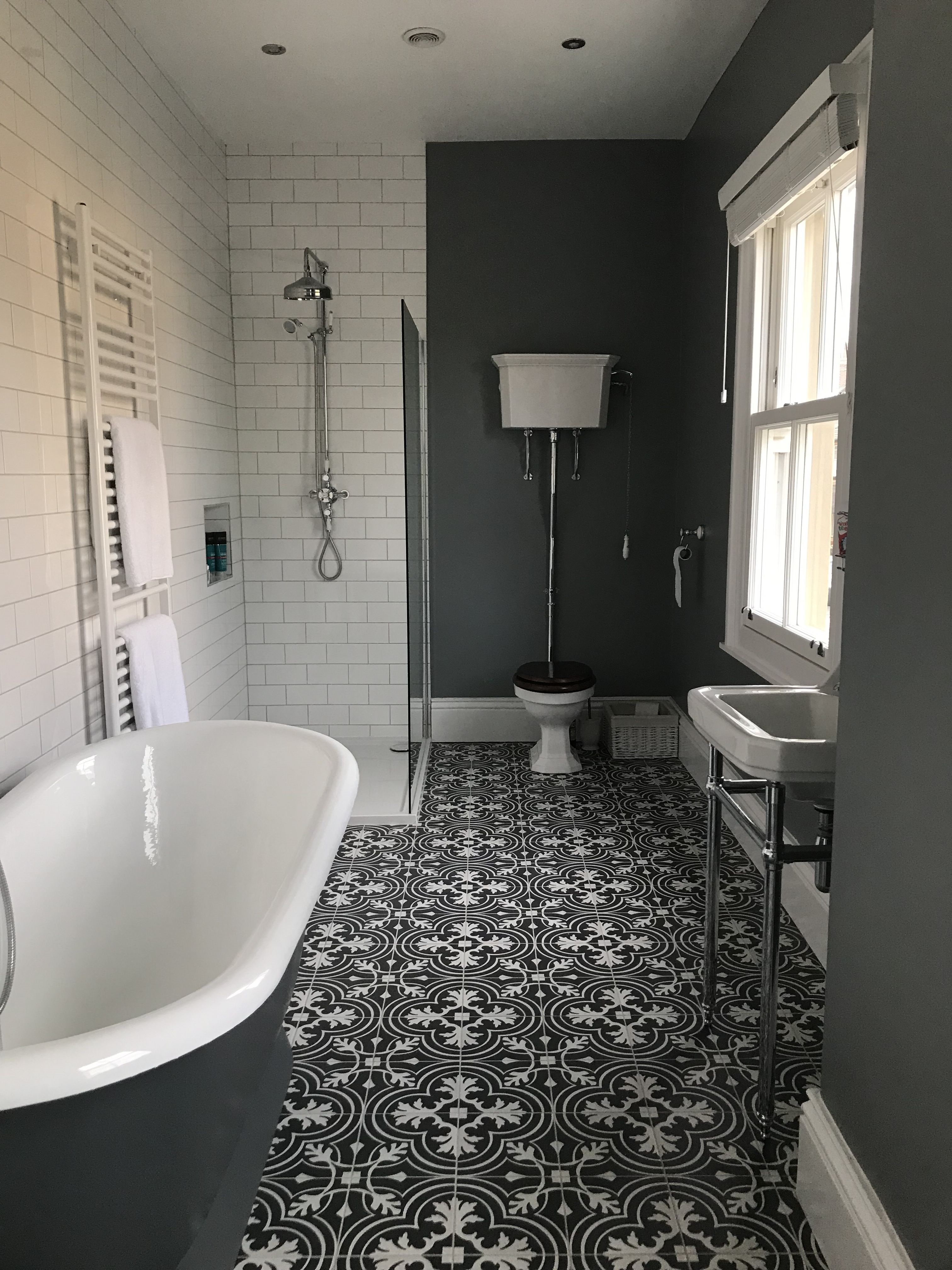 Product Lifestyle image of a Victorian style bathroom, with black and white patterned tiles, a roll top freestanding bath, an exposed pipework shower with riser rail and handset shower, a chrome washstamd with a stepped washbasin, an open back toilet with a black seat and a high level cistern