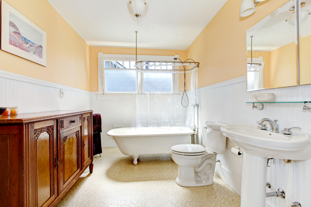 Lifestyle image of a vintage style bathroom, featuring a roll top showerbath with painted white clawfeet and a shower curtain, an open back close coupled toilet, a pedestal basin, a glass shelf, a mirror cabinet and a wooden bathroom cabinet#