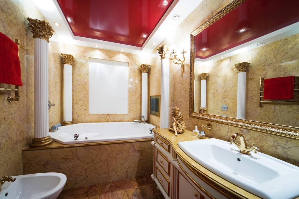 Lifestyle image of a white and gold bathroom design, featuring a built in jacuzzi bath with four white and gold pillars in each of its corners, gold marble walls, gold framed mirror, gold radiator and a white washbasin unit with gold accenting around its drawers and doors