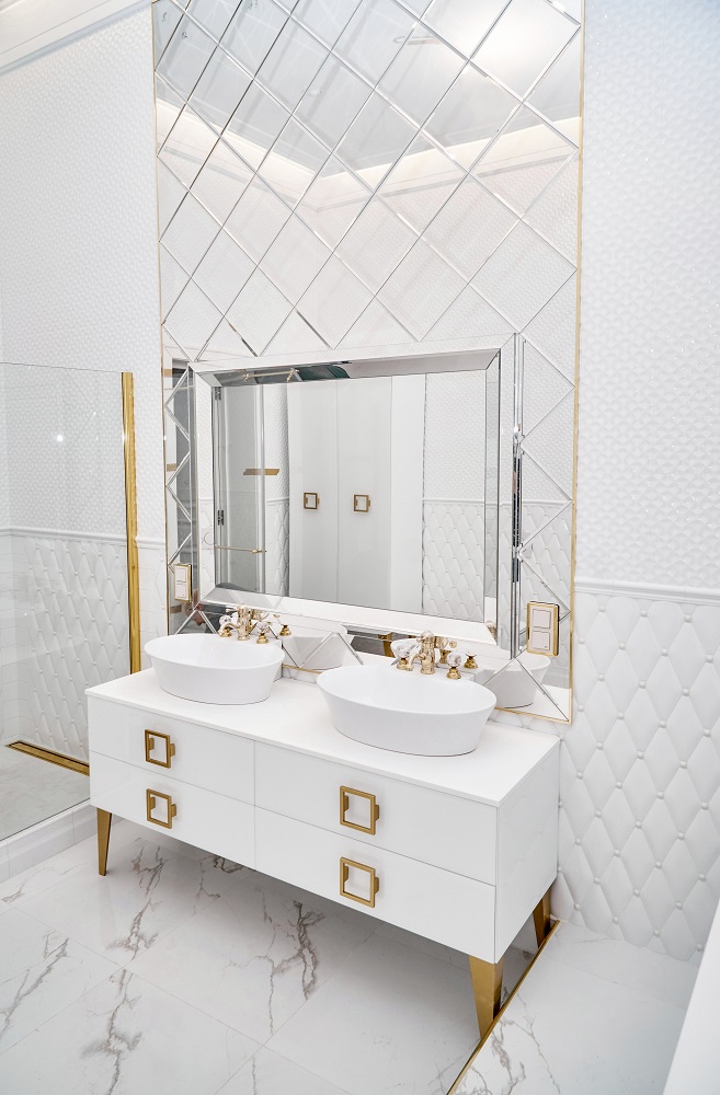 Lifestyle image of a white and gold bathroom design, featuring a white washbasin unit with gold feet and handles, countertop basins with gold taps, a mirror with gold trim and shower screens with gold brackets