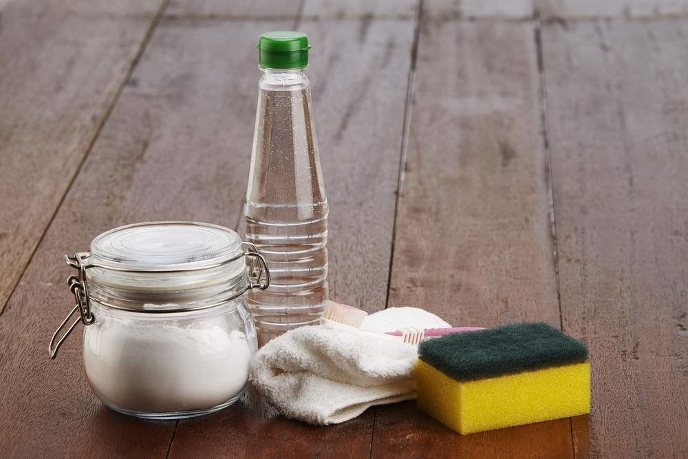 Close up image of a jar of bicarbonate of soda, white vinegar, a cloth, a toothbrush and a sponge