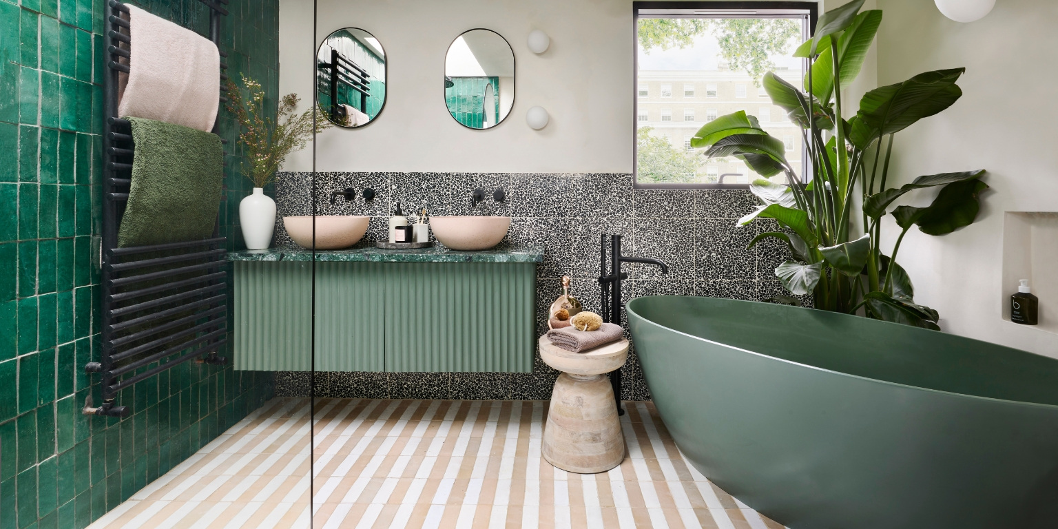 Bathroom with a green freestanding bath, fluted sage vanity with twin pink basins, green wall tiles, a terrazzo splashback and a large leafy plant beside the window
