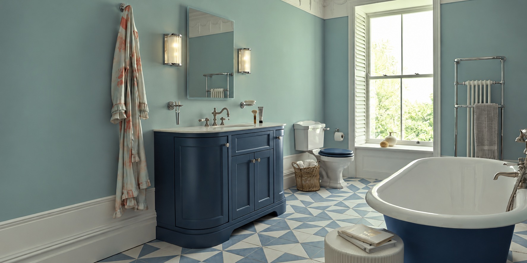 A stylish blue traditional bathroom featuring a freestanding bath, navy vanity unit, patterned floor tiles and soft natural light from a large window.