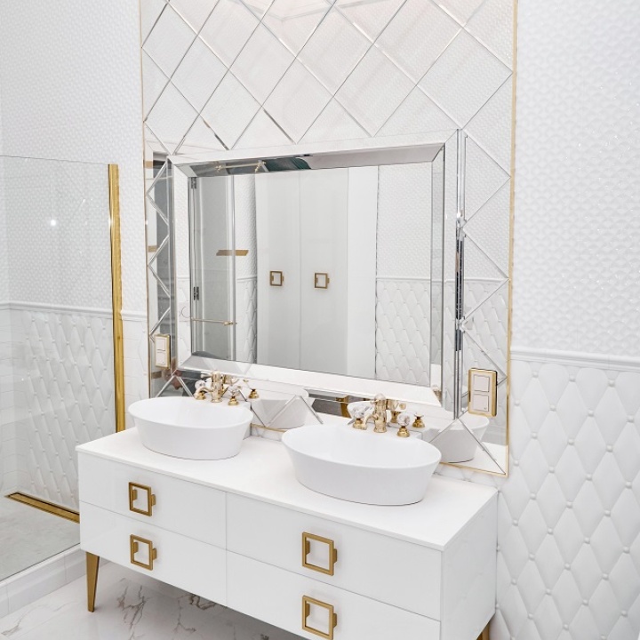 Lifestyle image of a white and gold bathroom, with a painted white double washbasin unit with gold feet and handles, gold taps and a gold bracket on the glass shower screen