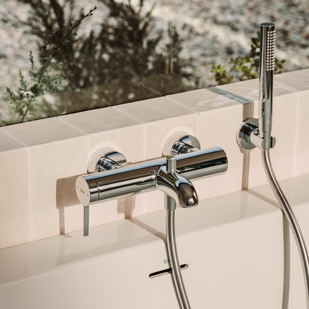 Lifestyle image of the Roca Ona Chrome Wall Mounted Bath Shower Mixer mounted on a bathroom wall and above a bathtub. Resting next to a microphone style handset shower.