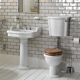image of a traditional toilet and basin set. It features a low level toilet with a chrome pipe and brown wooden seat, with a white pedestal basin with backsplash design, chrome accessories and mirror on a light metro tiled walll