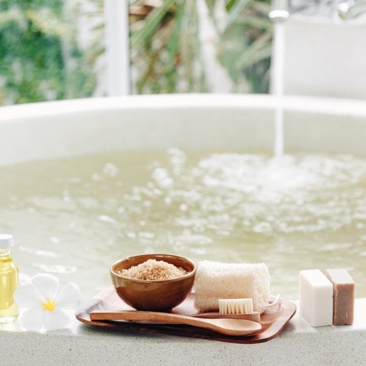 Lifestyle image of a tray on the edge of a bath, holding a sponge, bathing brush and bath salts, as well bars of soap and bottles of body wash