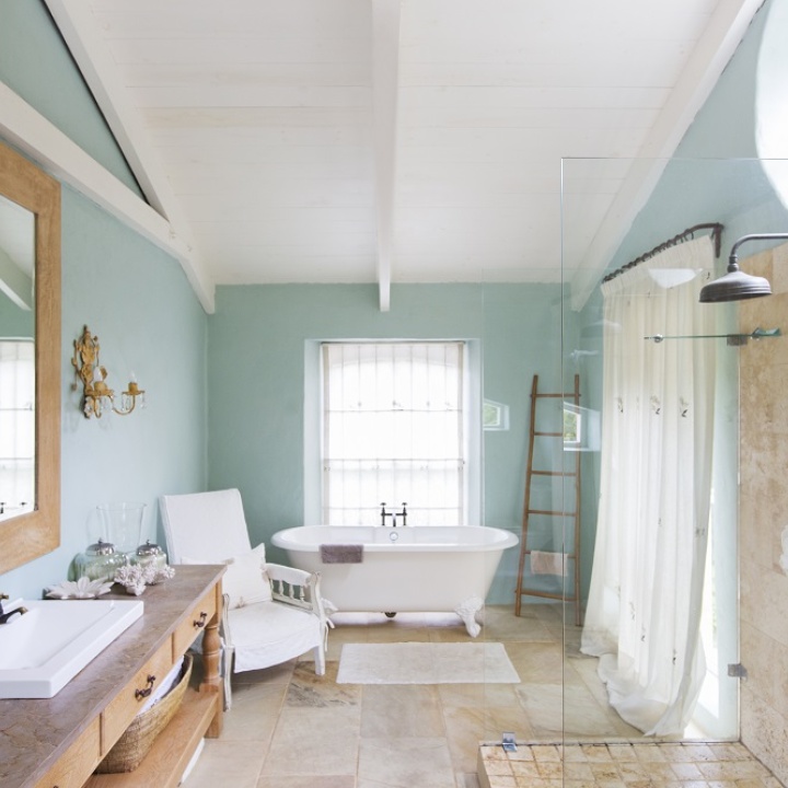 Lifestyle image of a farmhouse style bathroom, with wooden floors and turquoise walls, a walk in shower enclosure, wooden washstand, white freestanding bath with painted legs and white curtains across the windows